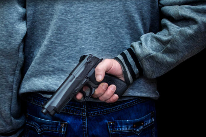 Man,Holding,A,Gun,Behind,His,Back,Against,A,Black