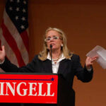 Woman speaking at podium with "DINGELL" sign.
