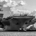 Legendary Carrier RETIRES—Navy Scrambles With No Backup Large aircraft carrier being assisted by tugboats in the ocean
