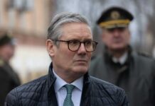 A man in formal attire with glasses, looking serious at an outdoor event