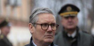A man in formal attire with glasses, looking serious at an outdoor event