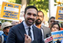 A man speaking at a rally with supporters holding protest signs