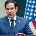 Green Cards Revoked—Rubio Drops The Hammer A man in a blue suit speaking into a microphone with an American flag in the background