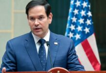 Green Cards Revoked—Rubio Drops The Hammer A man in a blue suit speaking into a microphone with an American flag in the background