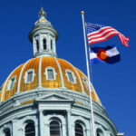 The dome of a state capitol building with flags flying against a blue sky