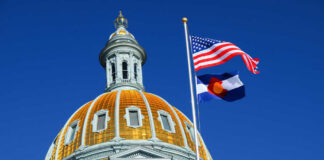 The dome of a state capitol building with flags flying against a blue sky