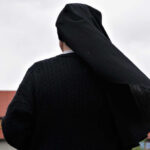 A nun in black attire standing outdoors with a cloudy sky in the background