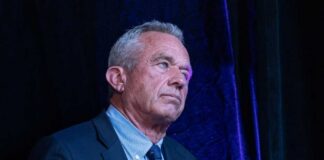 A man in a suit with a serious expression, illuminated by stage lighting