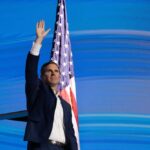 A man waving his hand in front of an American flag at a public event