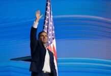 A man waving his hand in front of an American flag at a public event