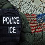 Police officer in tactical gear standing near a barbed wire fence with an American flag in the background