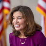 Hochul’s Anti-Trump Jab Backfires Spectacularly Smiling woman in a purple dress with flags behind