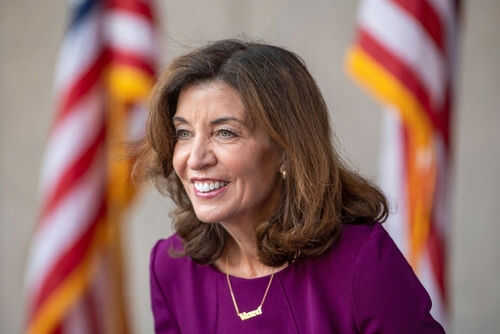 kathy-hochul2031652052 Smiling woman in a purple dress with flags behind
