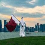 A person in traditional attire holding the Qatar flag against a city skyline at sunset