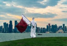 A person in traditional attire holding the Qatar flag against a city skyline at sunset