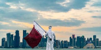A person in traditional attire holding the Qatar flag against a city skyline at sunset