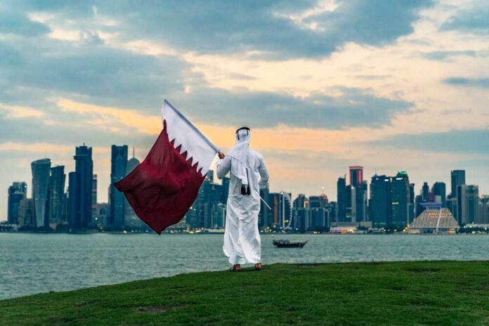 A person in traditional attire holding the Qatar flag against a city skyline at sunset