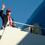 A man in a suit and red cap waving from the steps of an airplane