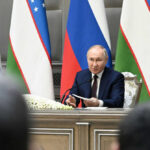 man in suit speaking at a conference table with microphones and flags behind