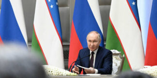 man in suit speaking at a conference table with microphones and flags behind