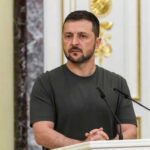 Zelenskyy Sounds Alarm, Then Nudges Peace Man speaking at podium with microphones, Ukrainian flag behind.