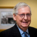 Power Vacuum Brewing? McConnell Hospitalized—Again Smiling man in suit with round glasses indoors.