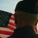 Disabled Vets’ $10K Raise Funded by Higher VA Loan Fees A veteran in uniform standing in front of an American flag at sunset