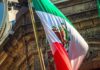 Mexico Targets ICE With Homicide Claims Mexican flag waving in front of an ornate stone building