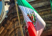 Mexico Targets ICE With Homicide Claims Mexican flag waving in front of an ornate stone building