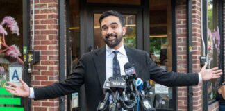 Man in a suit speaking at outdoor press conference with multiple microphones