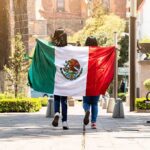 Fentanyl Fallout: Governor’s Return Sparks Uproar two people walking with a Mexican flag on a sunny city street