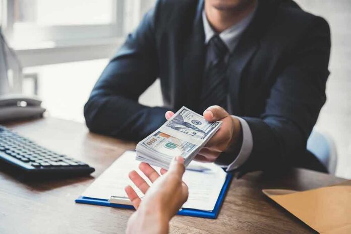 shutterstock_593433602 Businessperson hands over a stack of US dollars across a desk