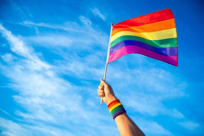 shutterstock_1089826724 Hand holding a rainbow pride flag against a blue sky