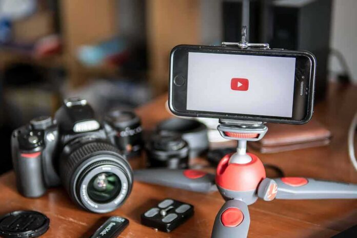shutterstock_1459193108 Smartphone on mini tripod beside DSLR camera and lenses