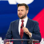 Michigan Erupts – Vance Torches ‘Weasel’ Rival man in suit speaking at a podium