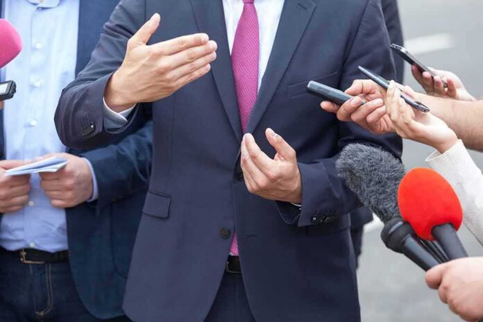 shutterstock_1040735278-1 Businessperson in suit speaking to reporters with microphones