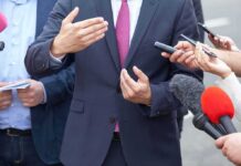 Businessperson in suit speaking to reporters with microphones