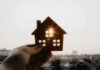 Hand holding a small house cutout against a bright sunset sky