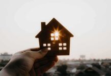 Nonstop Fees – Homeownership’s Hidden Bill Explodes Hand holding a small house cutout against a bright sunset sky