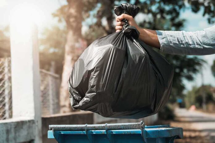shutterstock_1267099906 Hand dropping a black garbage bag into a blue bin