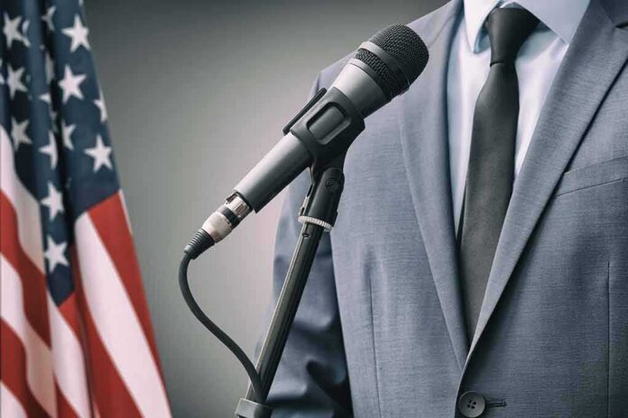 shutterstock_1614364942 Microphone at podium with person in suit and US flag