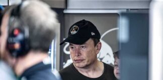 Elon Musk wearing a black cap in a crowded indoor setting